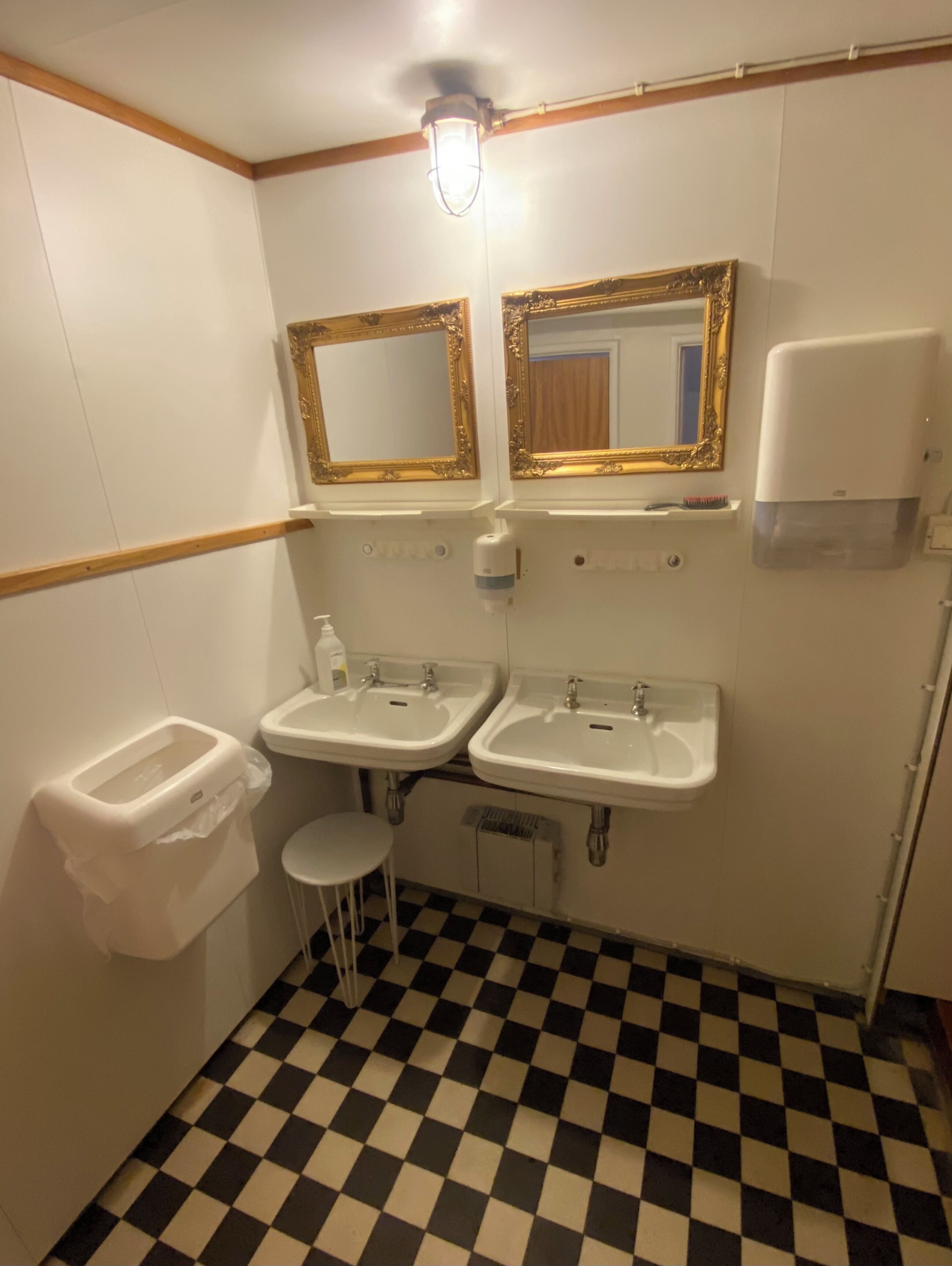 A bathroom with two sinks, a toilet, a small stool, and a mirror with an ornate frame.