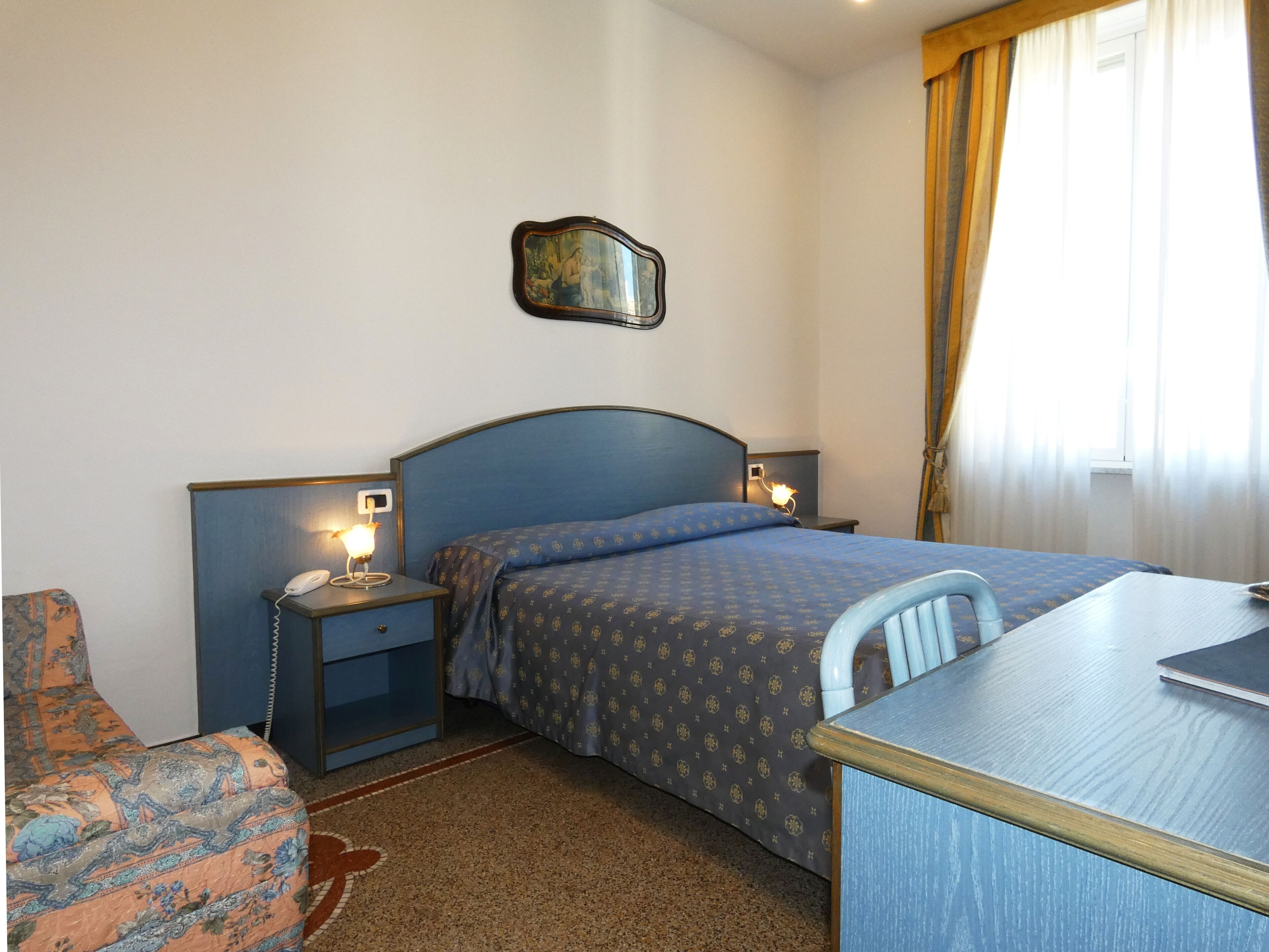Quarto de hotel com cama, escrivaninha, cadeira, mesa de cabeceira com telefone e espelho fixado na parede.