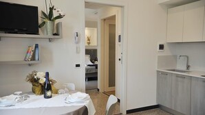 Quarto de hotel com mesa de jantar posta para dois, televisão fixada na parede, pequena área de cozinha com pia e porta que dá para um quarto.