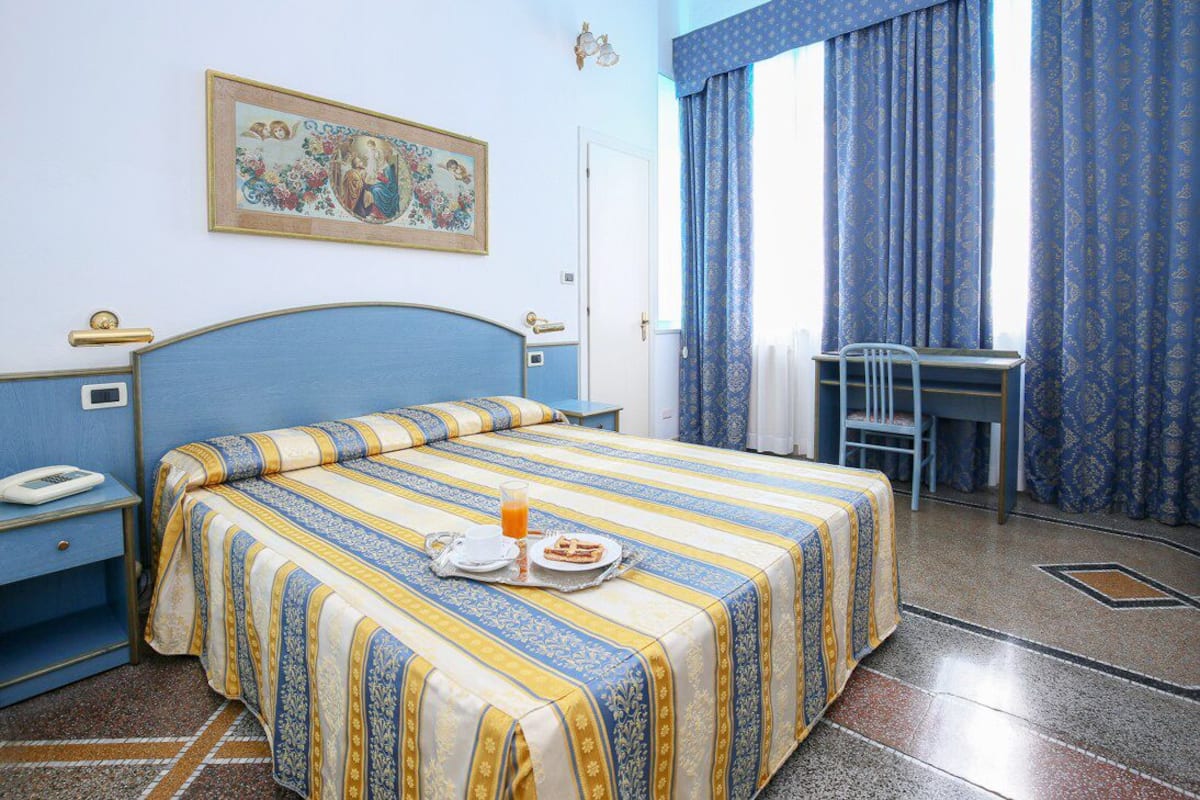 A hotel room with a bed, a desk, a chair, a telephone, and a painting on the wall.
