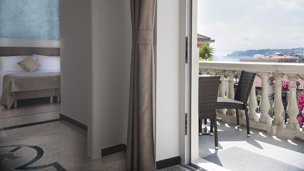 Quarto de hotel com cama, vista para o mar e varanda com cadeiras de vime.