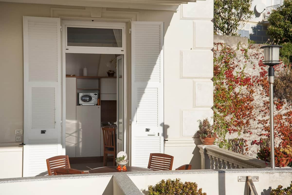 A balcony with a view of a garden, a door leading to a room with a microwave, and a chair.