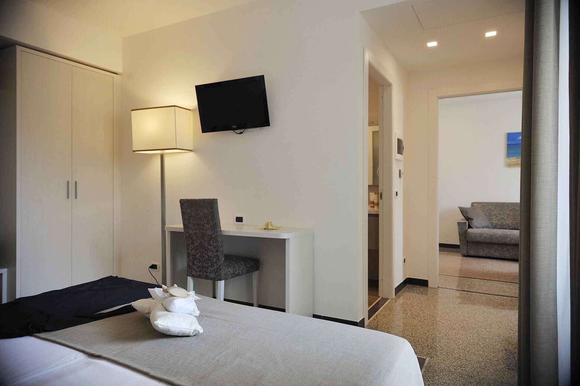 Ein Hotelzimmer mit einem Bett, einem Schreibtisch, einem Sessel und einem Fernseher. Im anliegenden Raum befindet sich eine Couch.