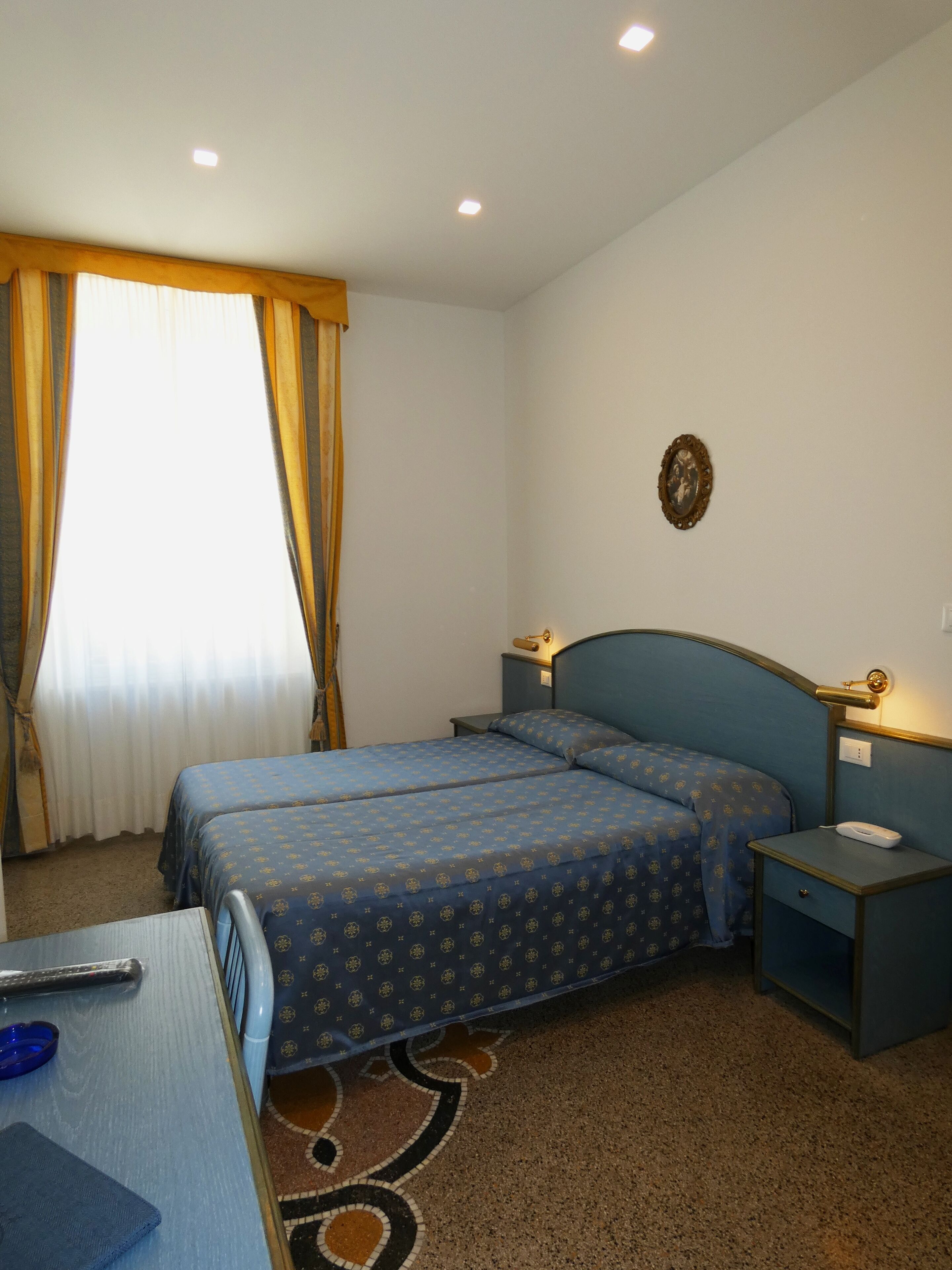 A hotel room with two beds, a desk, a chair, a nightstand, and a wall-mounted lamp.
