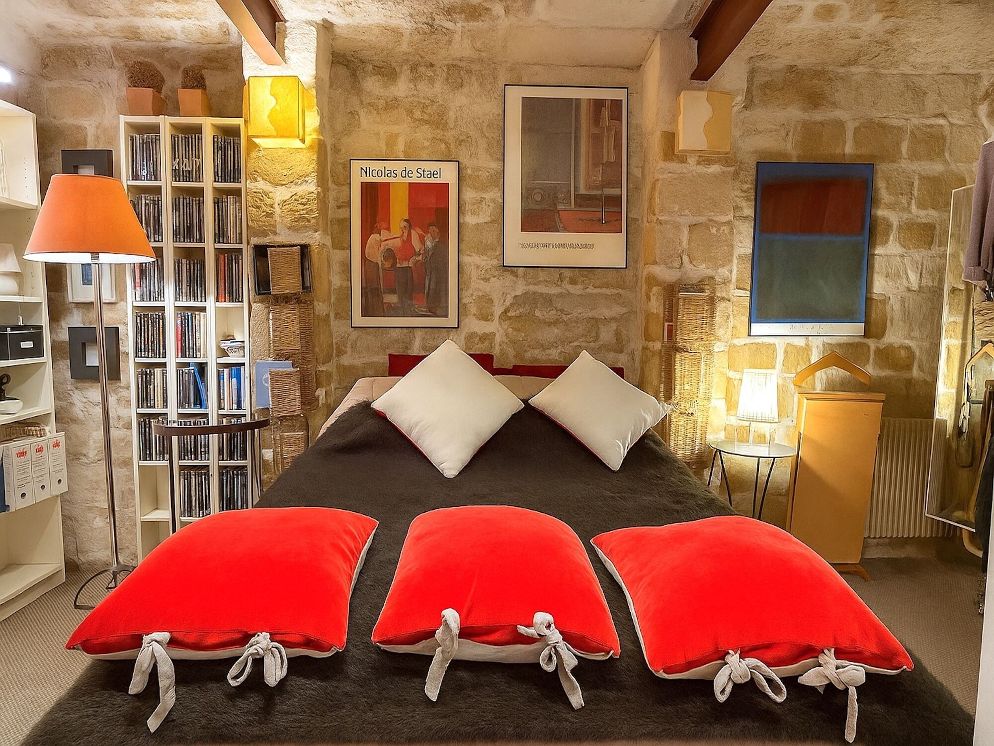 Ein Schlafzimmer mit Steinsturz, Bücherregal, Bett mit roten Kissen und einem gerahmten Bild an der Wand.