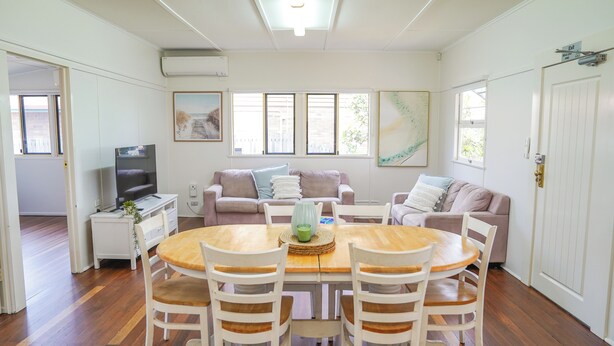 A spacious living room with a wooden dining table surrounded by white chairs, a beige sofa set, and a television on a white stand. - Seeside Cottage Bargara | Beach 100m | Pet-Friendly | Sleeps 6 (Bargara)