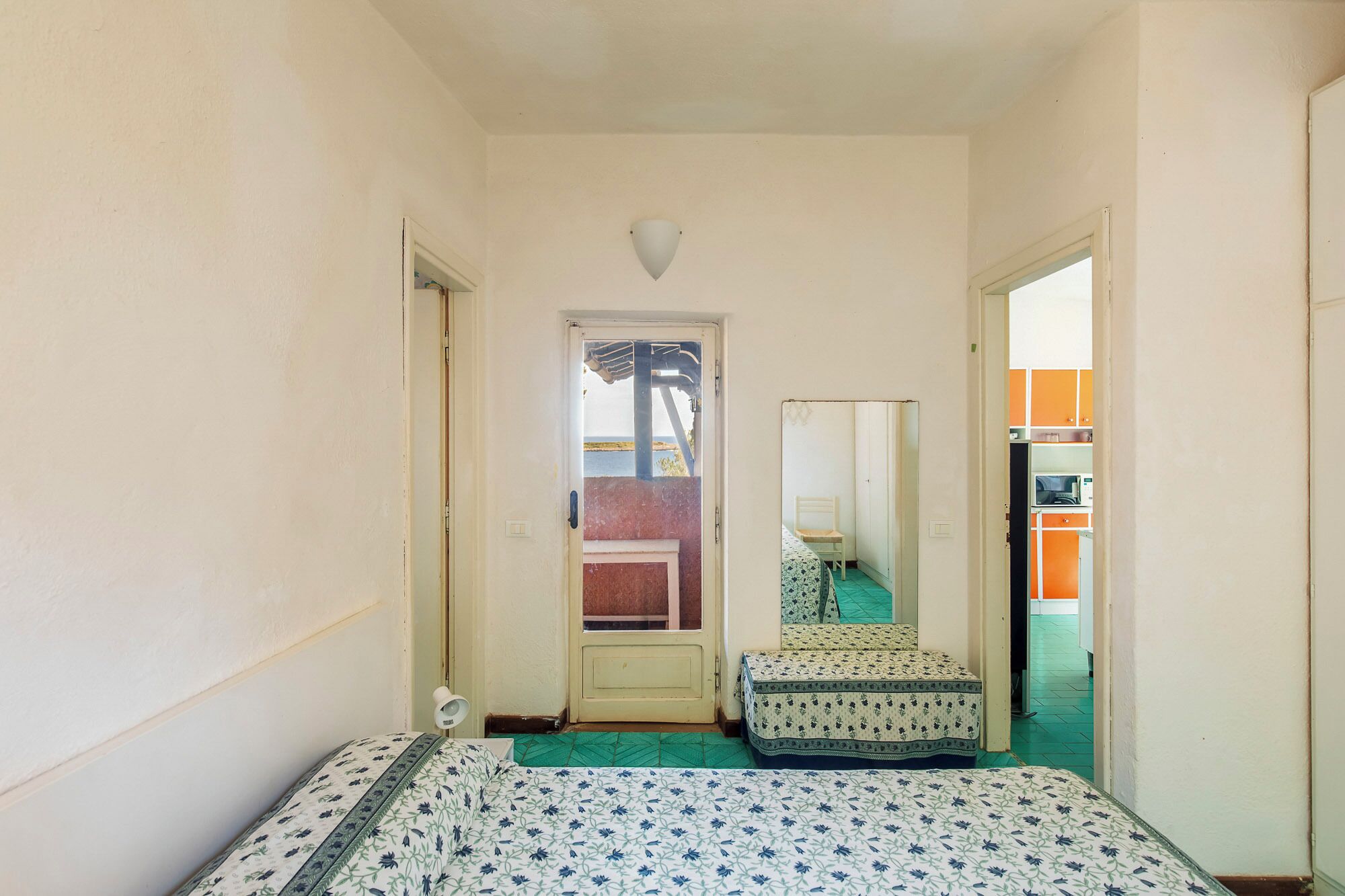 Un pasillo estrecho con una cama, una mesita pequeña y una silla. Se ve una puerta que da a una habitación con vistas al mar.