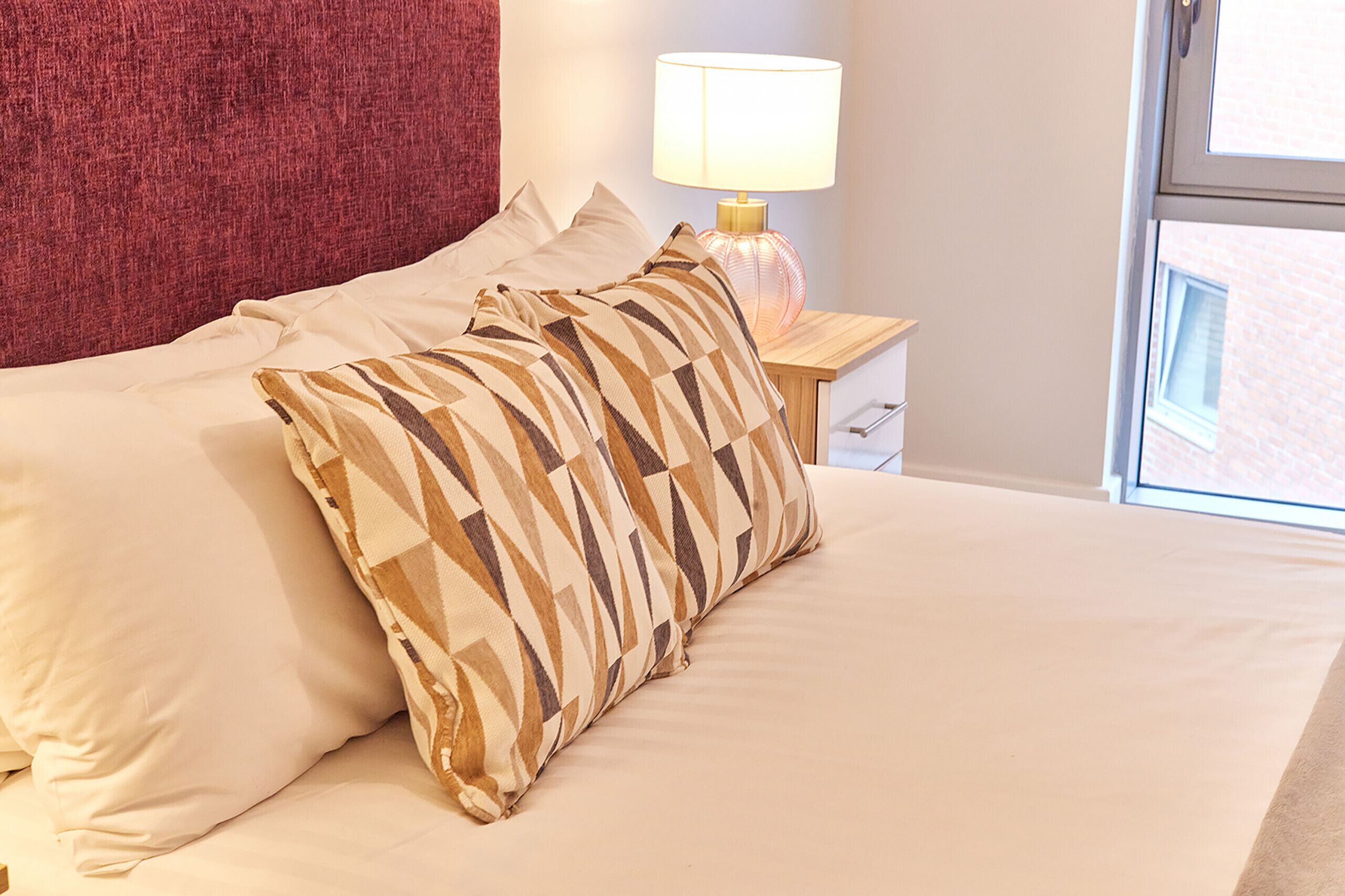 A neatly made bed with patterned pillows, a bedside lamp, and a window with a view of a building.