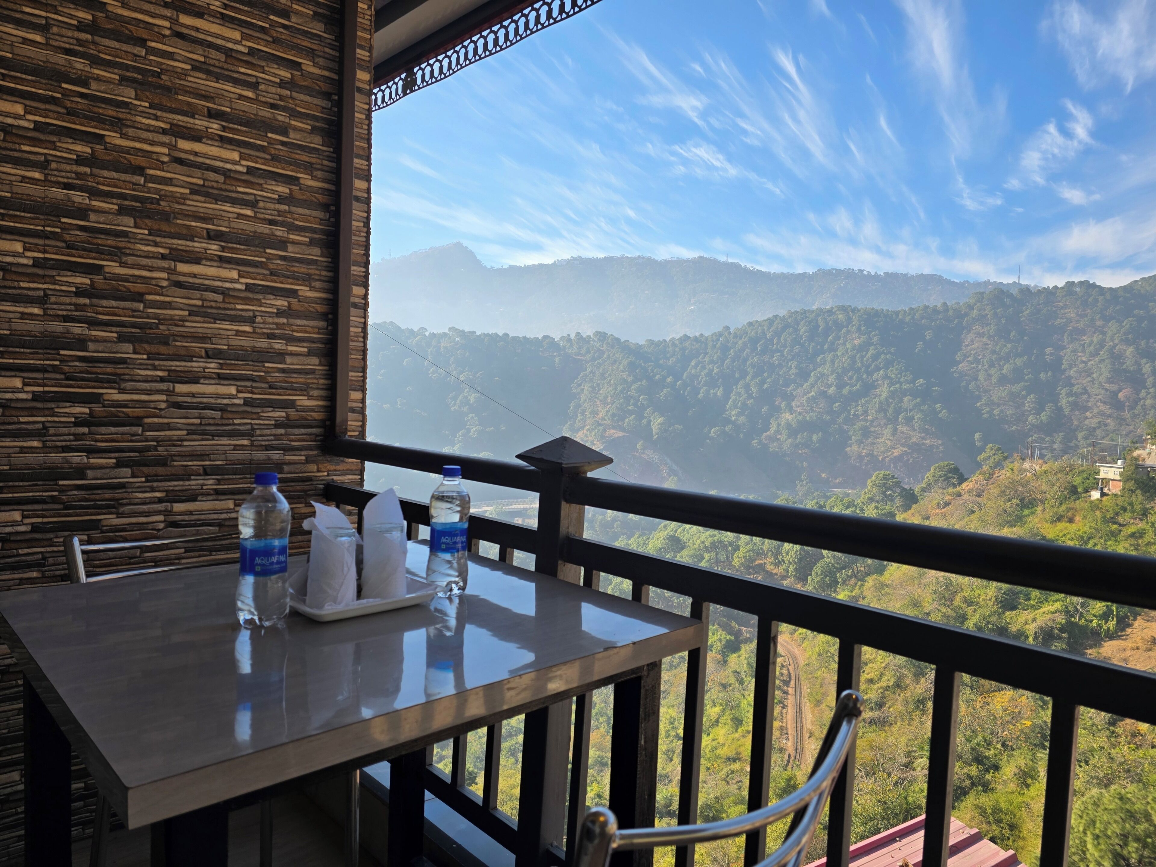 A balcony with a table, two water bottles, and a glass. The view includes a mountainous landscape and a clear sky.