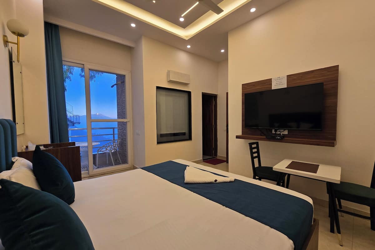A hotel room with a bed, a desk, a chair, a TV, and a balcony with a view of a mountain.