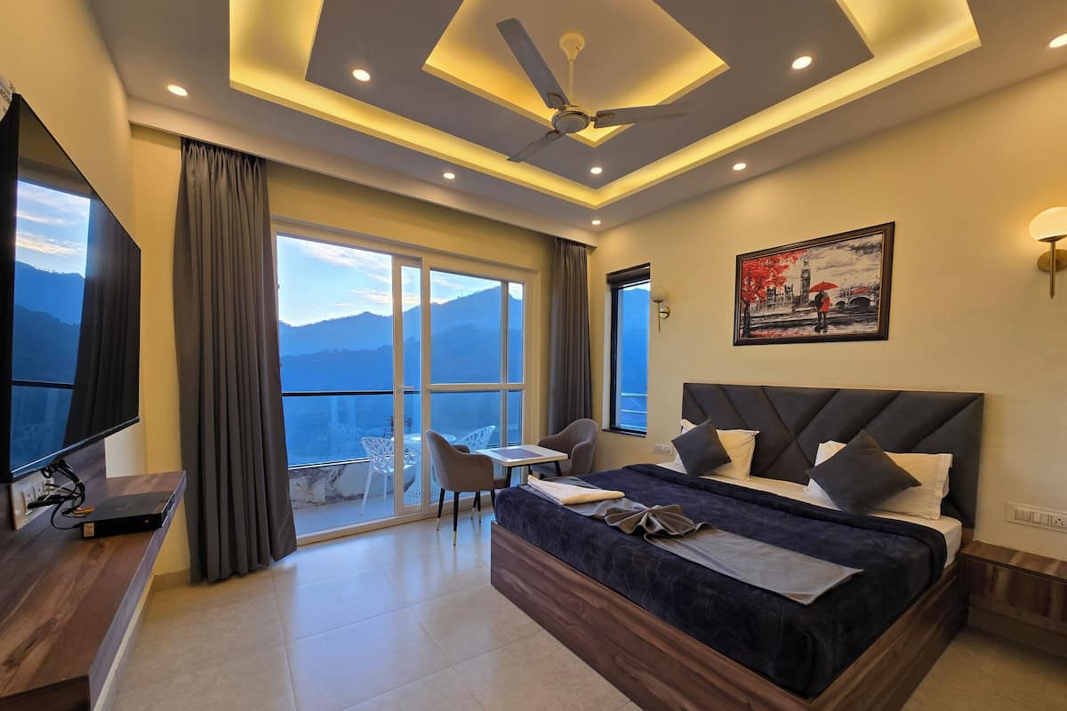 A modern hotel room with a large bed, a desk with a flat-screen TV, a balcony with a mountain view, and a ceiling fan.