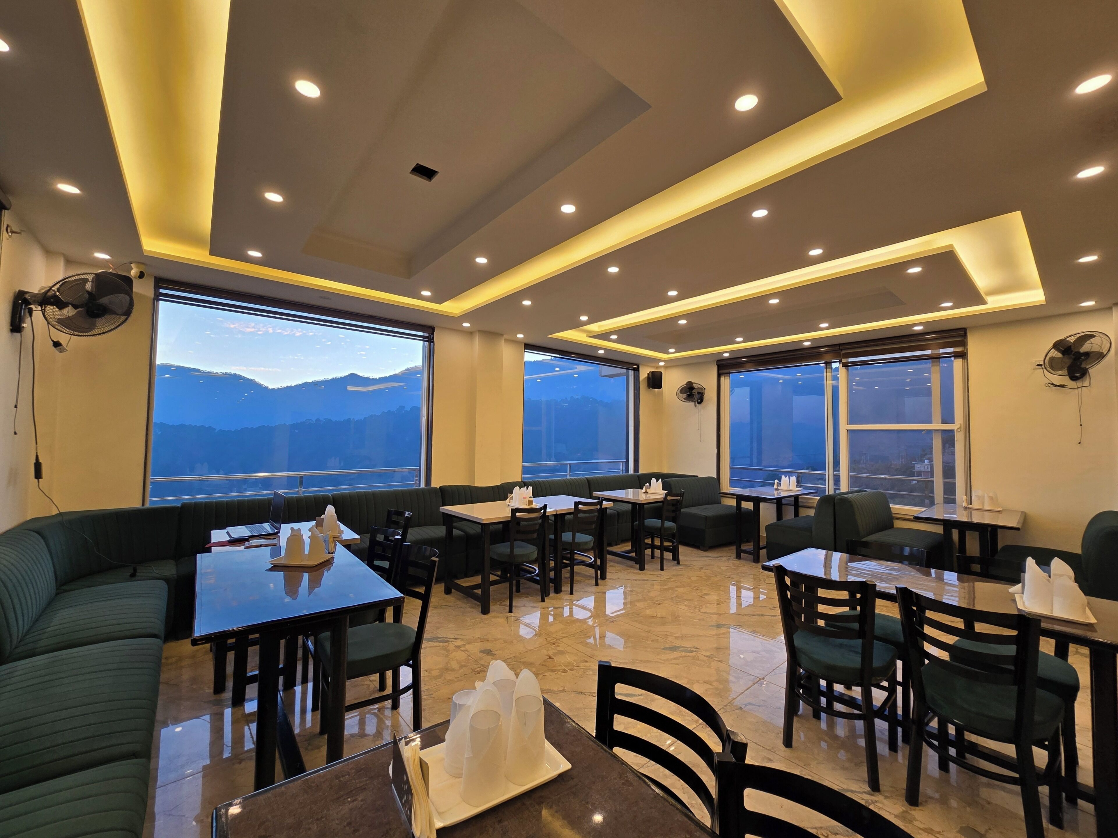 Un restaurant moderne offrant une vue sur les montagnes, avec des sièges, des tables et des ventilateurs de plafond de couleur verte.