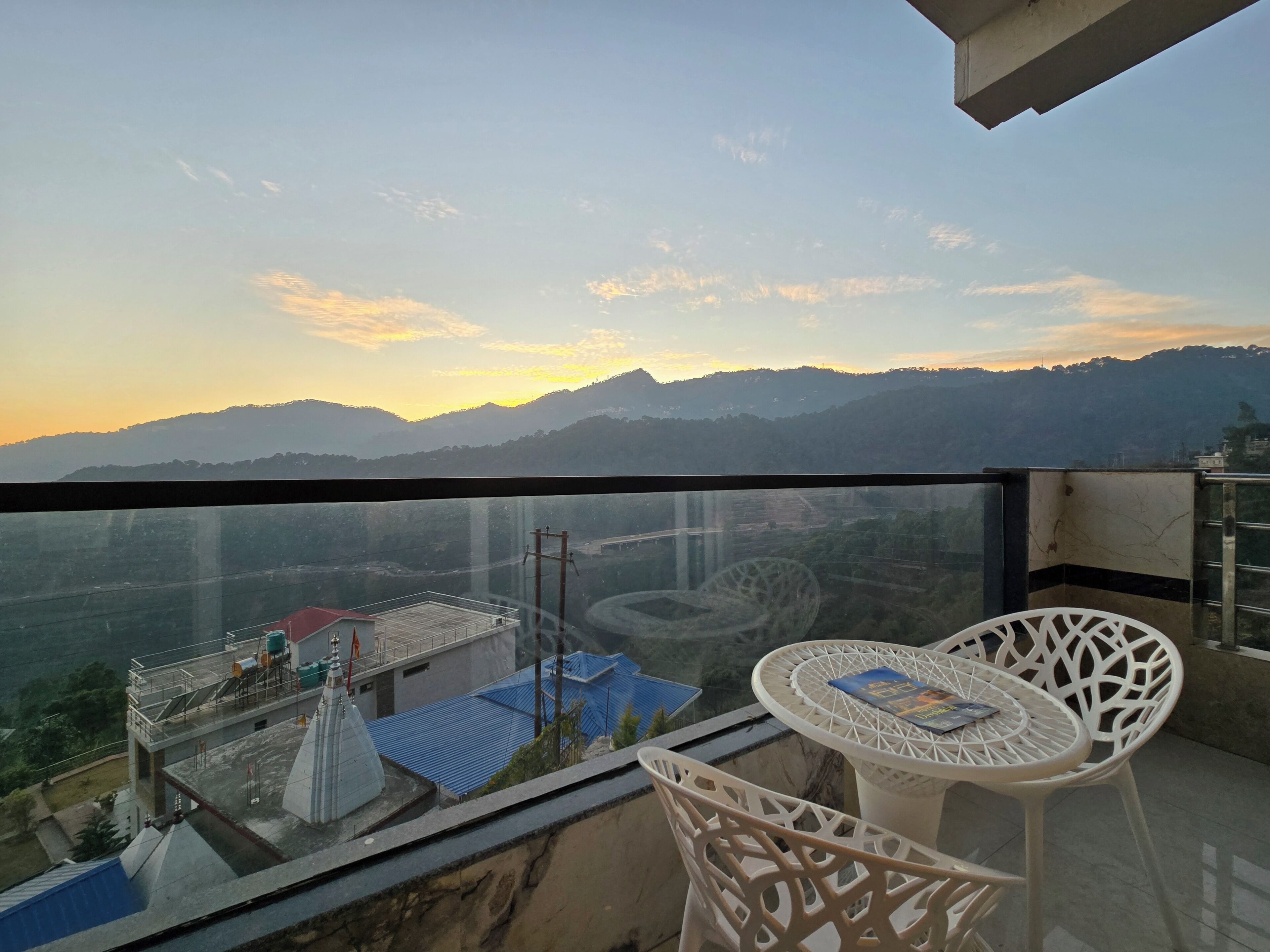 A balcony with a table and chairs, overlooking a mountainous landscape and a distant building with a blue roof.