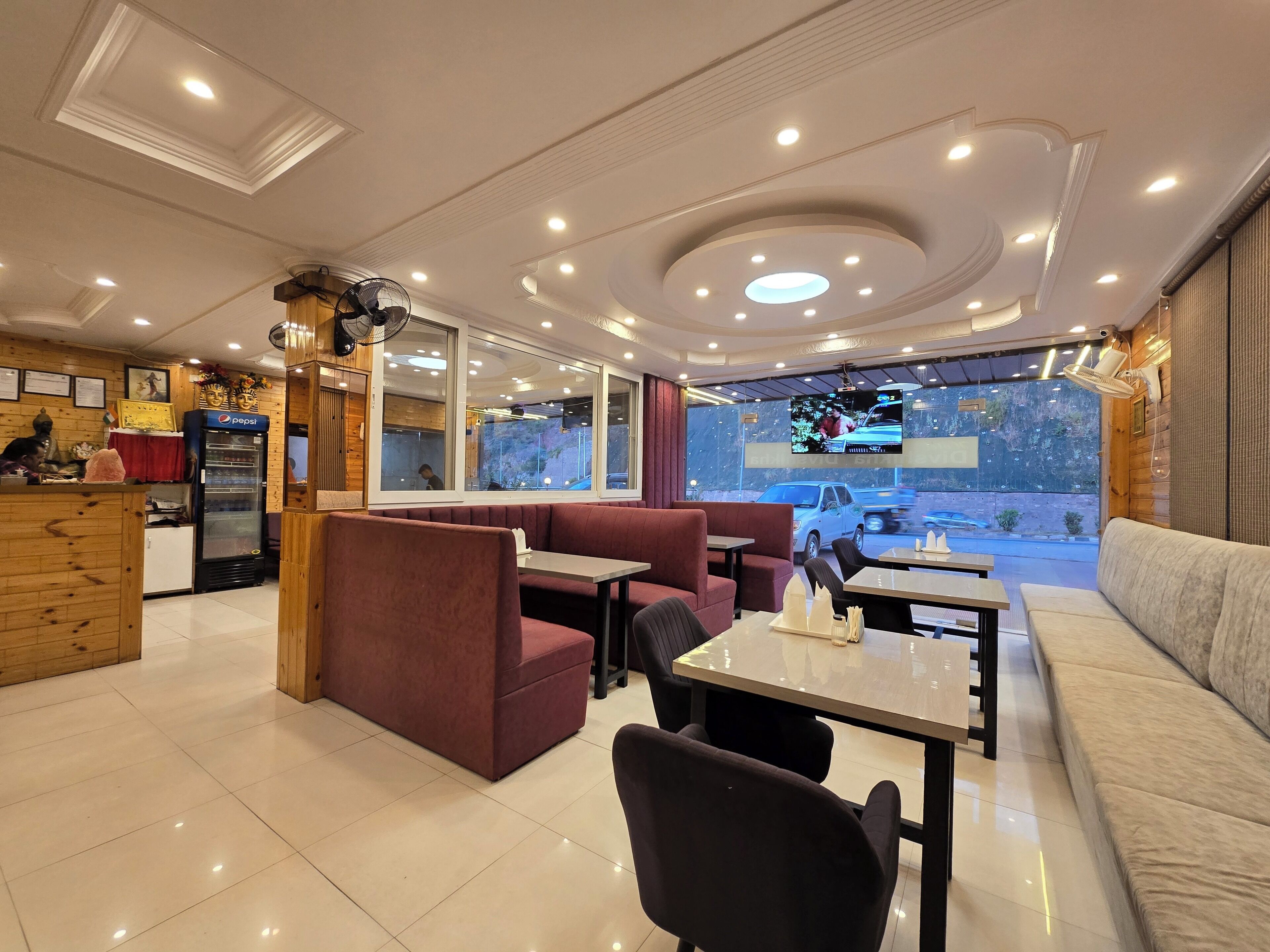A restaurant with a modern interior, featuring a seating area with red and beige couches, a counter with a Pepsi refrigerator, and a large window offering a view of the outside.