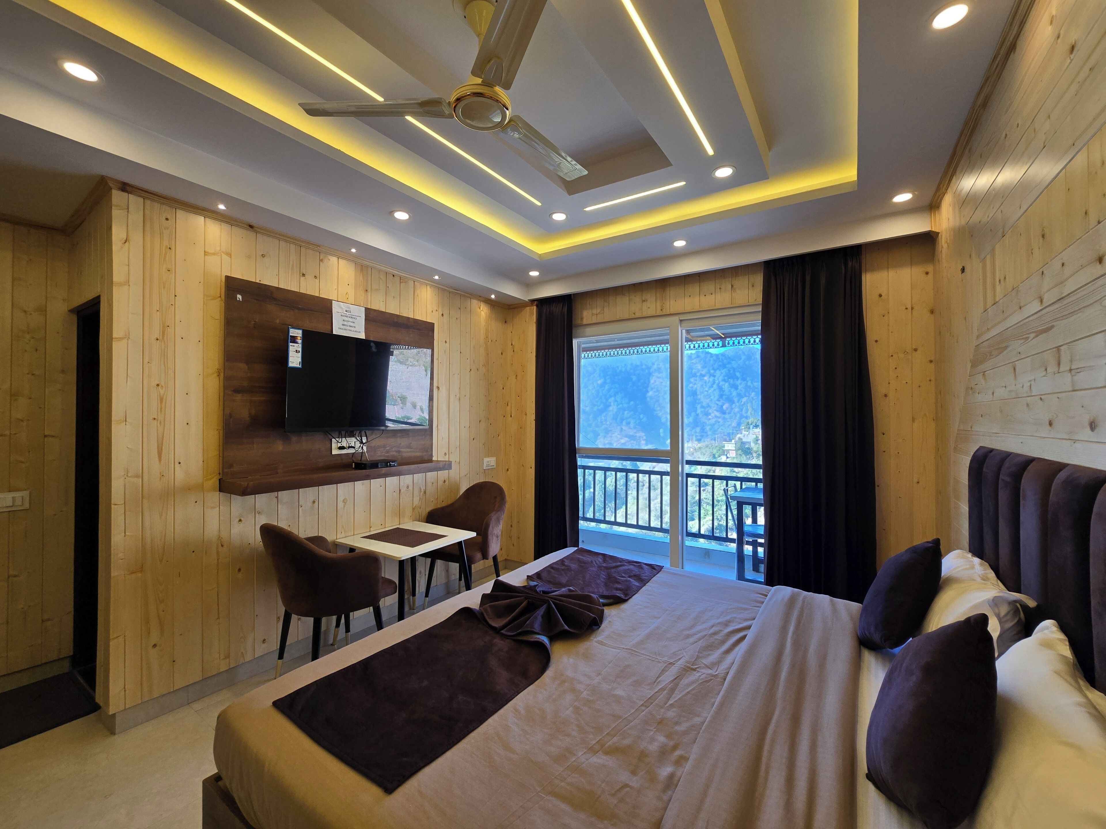 Ein Hotelzimmer mit Bett, einem an der Wand befestigten Fernseher, einem kleinen Tisch, zwei Stühlen und einem Balkon mit Bergblick.