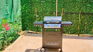 Een draagbare barbecue met twee branders en een bijbehorend plankje, geplaatst op een betonvloer met een groen gaashek en planten op de achtergrond.