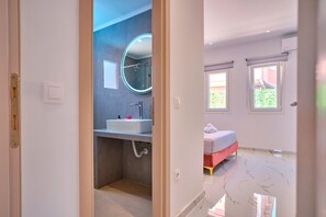 Ein modernes Badezimmer mit einem runden Spiegel, einem Waschbecken und einer Dusche. Der Raum verfügt über ein Bett mit rotem Kopfteil und ein Fenster.