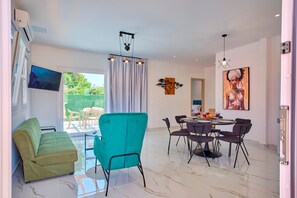 A modern living room with a green sofa, teal armchair, and dining area with a table set for six. - Drastis Escape Villas - Villa Eleni By Estia (Corfu)
