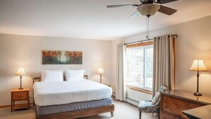 Ein ordentlich eingerichtetes Schlafzimmer mit einem Bett, zwei Nachttischlampen, einem Deckenventilator, einem Fenster mit Vorhängen und einem Bild an der Wand.