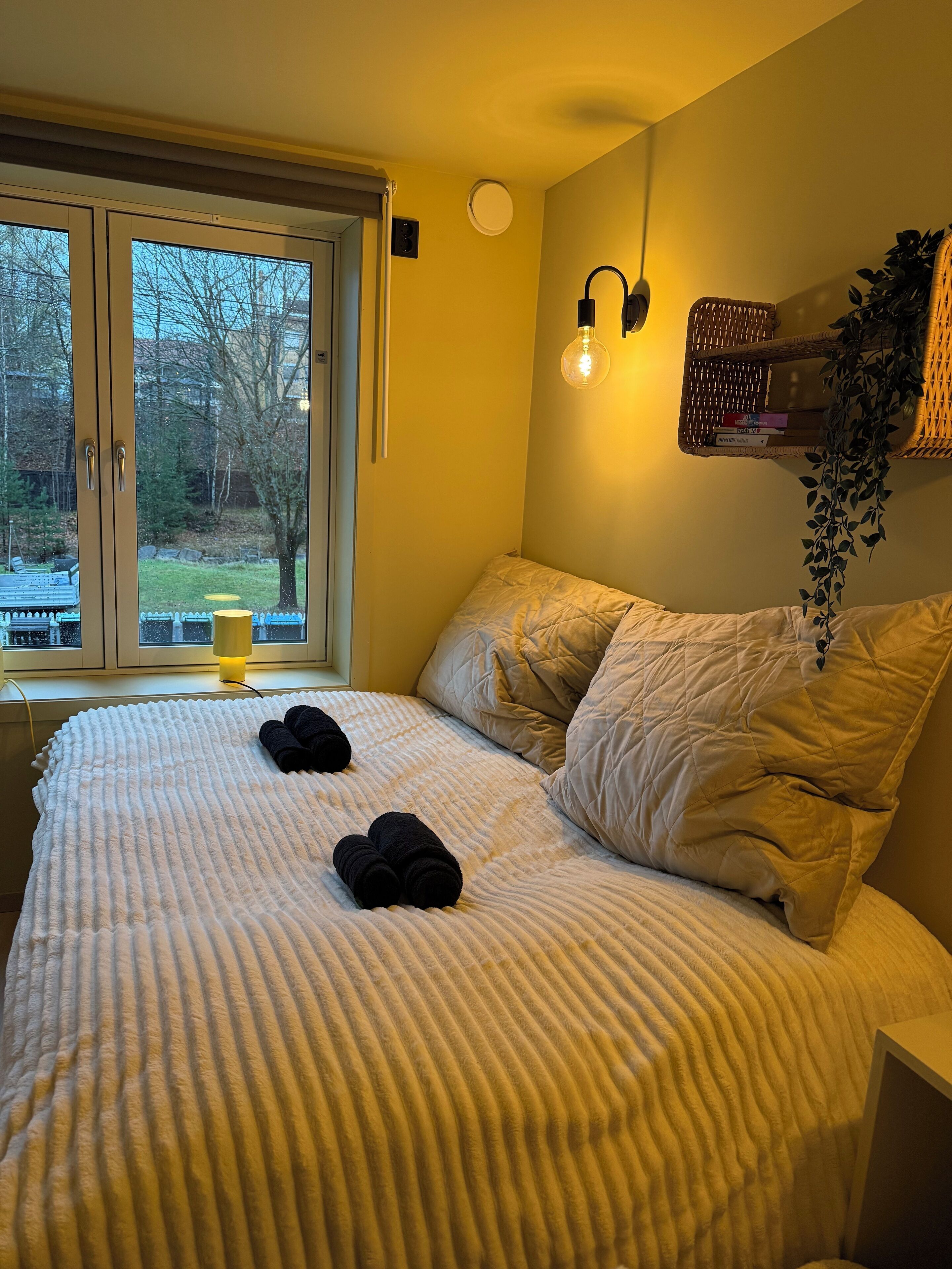 Een slaapkamer met een groot bed, een raam met uitzicht op bomen, een bedlamp en een speaker bevestigd aan de muur.