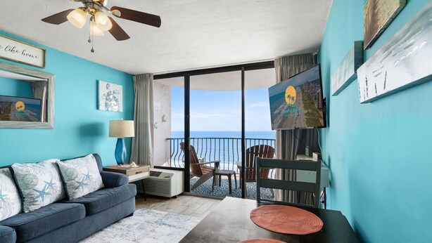 Ein Zimmer mit blauer Wand, Deckenventilator, Sofa mit Sternenmustern auf den Kissen, Tisch mit roten Untersetzern und Blick auf das Meer durch eine Schiebetür.