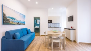 A modern living room with a blue sofa, a dining table with chairs, a kitchen area with a TV, and a painting on the wall. - Muzy Flat at Capuana by Napoliapartments (Naples)