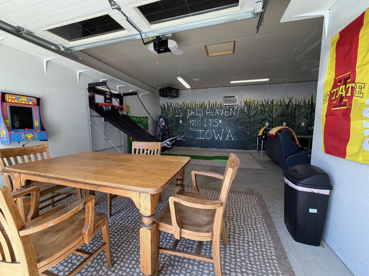 Une pièce avec une table et des chaises en bois, un tableau noir sur lequel est écrit : « EST-CE LE PARADIS ? NON, C’EST L’IOWA », un panier de basket et un drapeau sur lequel on peut lire « ÉTAT 2 ».