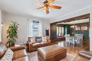 Ein geräumiges Wohnzimmer mit einer braunen Ledersofa, einem Essbereich mit weißen Stühlen und einem Deckenventilator.