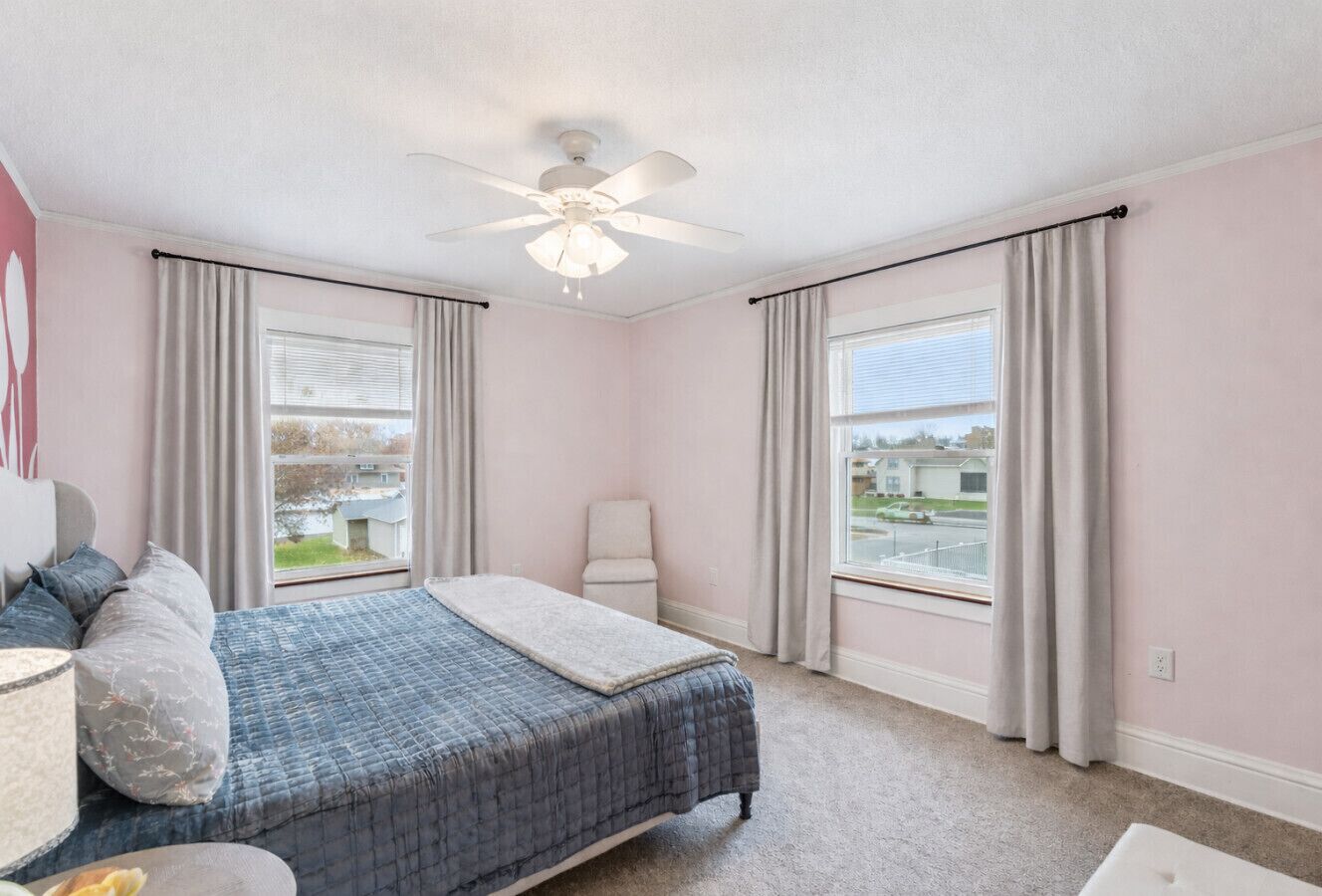 A bedroom with a bed, a chair, a desk, a ceiling fan, and large windows with curtains.