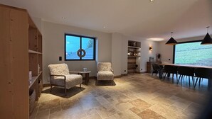 Amplia habitación con dos sillones cómodos, una estantería de madera, zona de comedor con una mesa larga y sillas, y un gran ventanal con vistas al exterior.