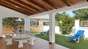 Eine Terrasse im Freien mit Tisch und Bänken, ein überdachter Bereich mit Holzboden und Blick auf ein Haus und Grünflächen.