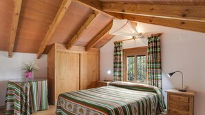 Ein Schlafzimmer mit Bett, Nachttisch, Lampe, einem Fenster mit Vorhängen, einer Zimmerpflanze und einem Holzschrank.