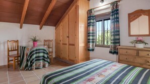 A room with a bed, wooden wardrobe, a table with a striped tablecloth, chairs, and a window with curtains. - Holiday Home 'Miguelin 8' with Shared Pool, Wi-Fi and Air Conditioning (Zahora)
