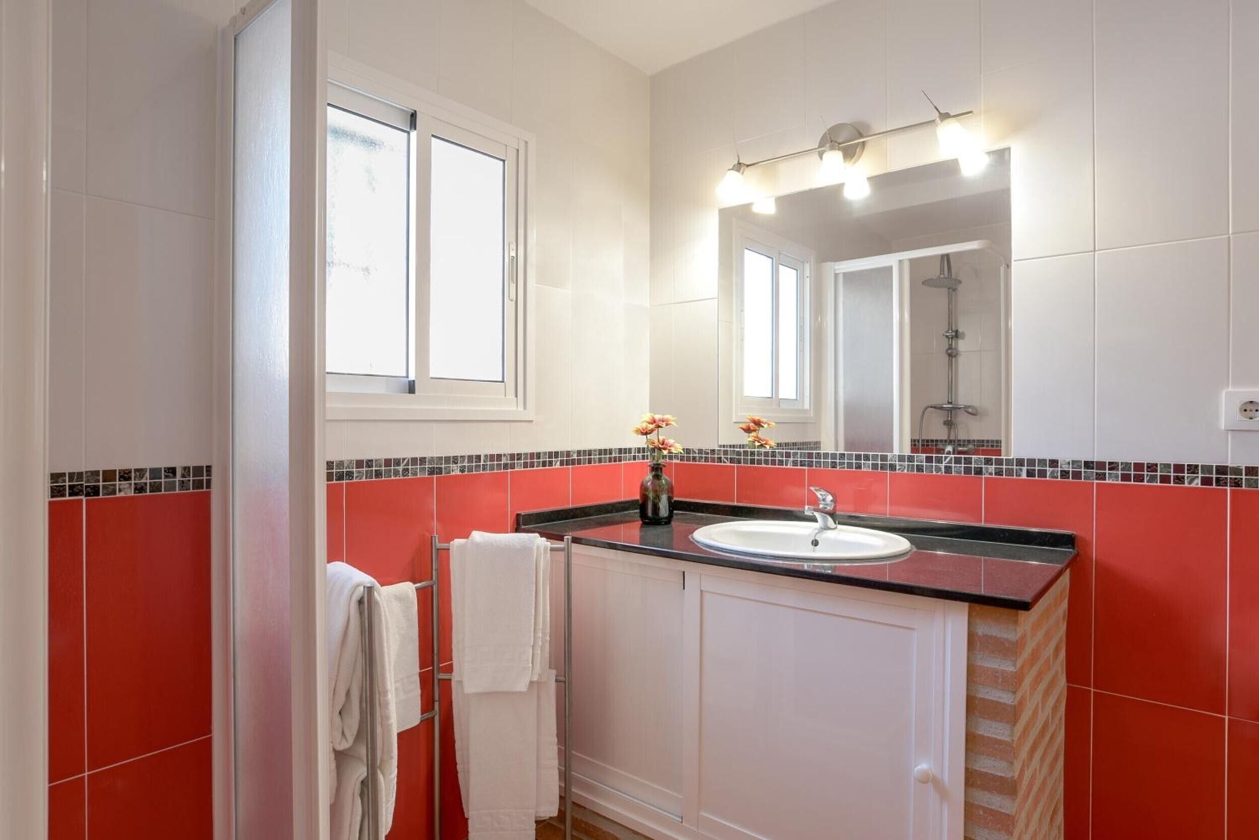 A bathroom with a white sink, red tiled walls, a mirror with lights, and a window.