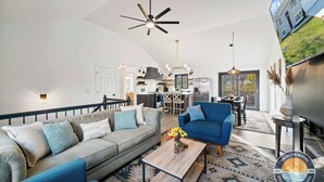 A spacious living room with a grey sofa, blue armchair, and a coffee table. A ceiling fan and pendant lights are visible. The kitchen is in the background with a dining area. - Lake Geneva House: Family Fun with Pool Table (Lake Geneva)