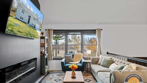 A modern living room with a flat-screen TV, a sofa, a coffee table, and a view of a house through large windows. - Lake Geneva House: Family Fun with Pool Table (Lake Geneva)