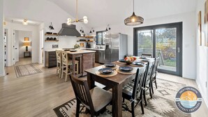A modern kitchen with a central island, dining area with wooden chairs, and a large window providing a view of the outdoors. - Lake Geneva House: Family Fun with Pool Table (Lake Geneva)