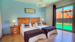 Ein Schlafzimmer mit zwei Betten, Holz-Kopfteilen, Nachttischlampen, einem gerahmten Kunstwerk und Blick auf eine grüne Außenanlage durch eine Schiebetür.