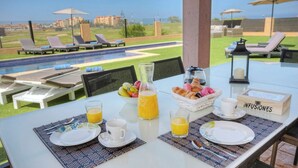 Ein Frühstückstisch mit Blick auf ein Schwimmbecken und Liegestühle, eine Orangensaftflasche, ein Korb mit Gebäck und ein Schild mit der Aufschrift 'INFUSIONES'.
