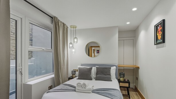 A modern hotel room with a large bed, a bedside table, a mirror, a window with curtains, and a wall-mounted picture. - Your London Escape | Stylish 3BR Apartment Stay (London)