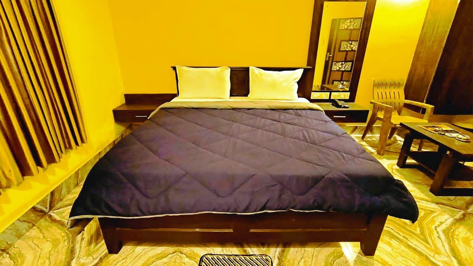 Cama com colcha escura, cabeceira de madeira e duas almofadas. Tem uma mesa de cabeceira de madeira com abajur, uma cadeira e uma pequena mesa com um livro e um telefone.