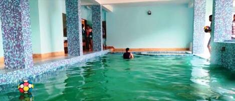 Piscina com azulejos de mosaico, uma pessoa nadando e vista do interior.