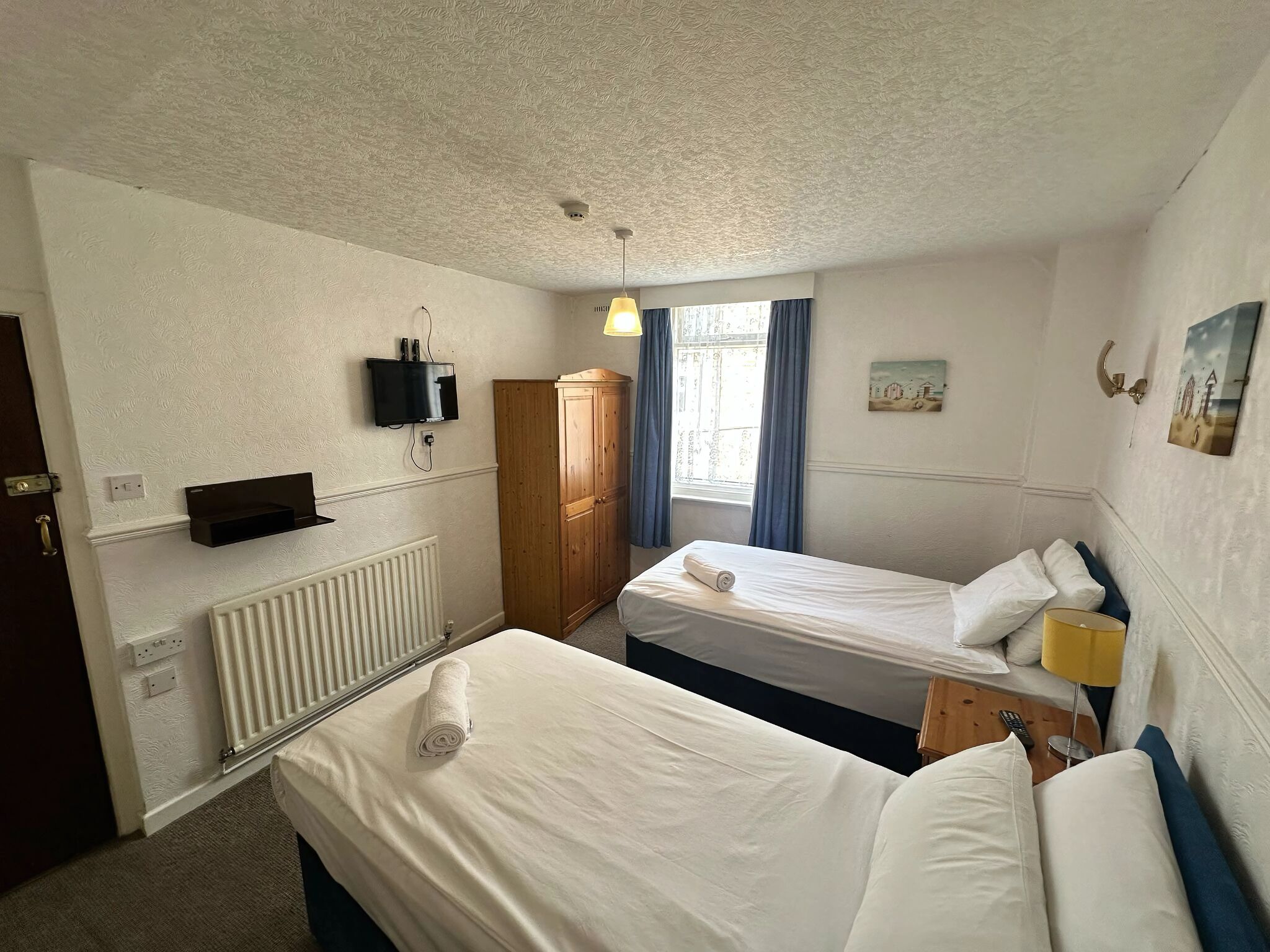 Habitación de hotel con dos camas individuales, un armario de madera, un televisor montado en la pared y una ventana con cortinas.