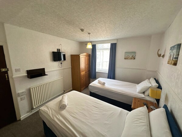 Habitación de hotel con dos camas individuales, un armario de madera, un televisor montado en la pared y una ventana con cortinas.