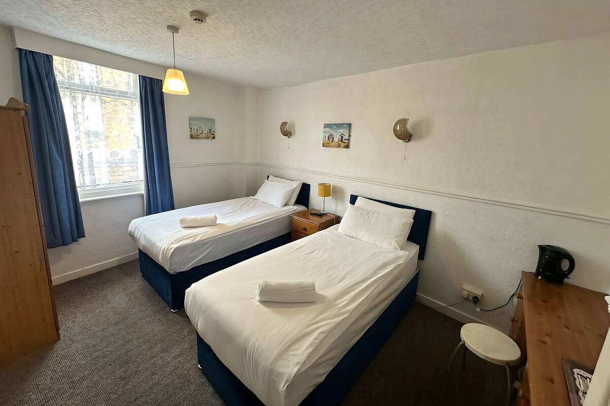 Habitación de hotel con dos camas, un escritorio, una silla, un taburete pequeño, una ventana con cortinas e iluminación en el techo.