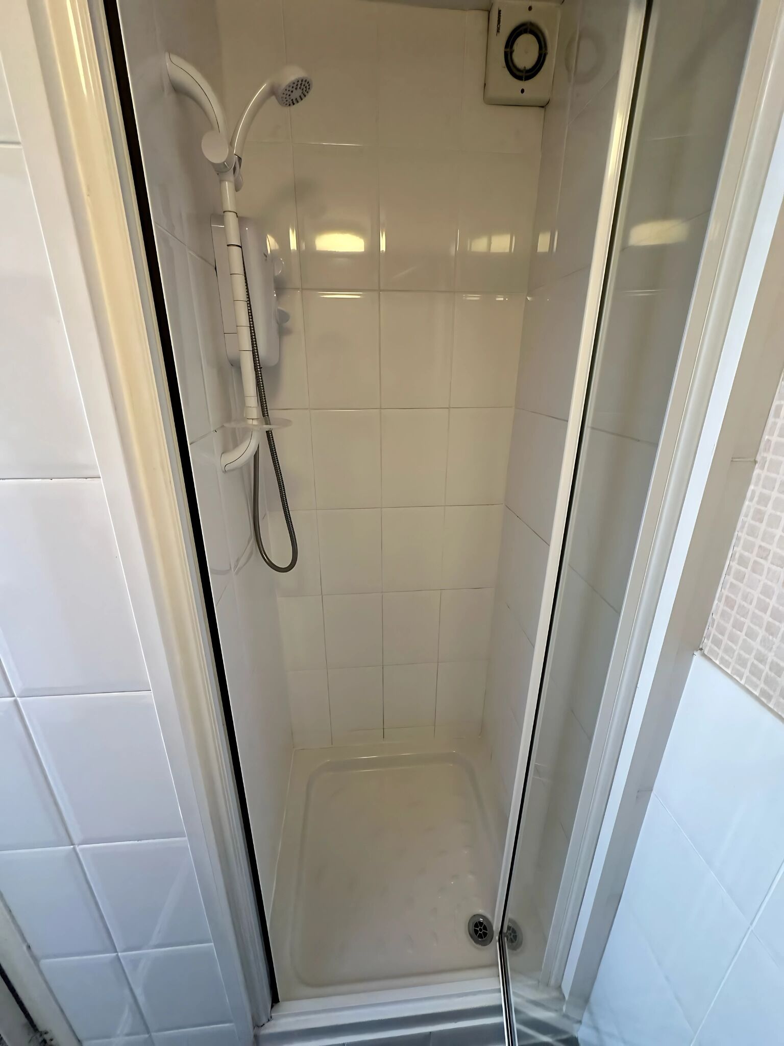 Ducha con mampara de vidrio, azulejos blancos, cabezal de ducha de mano y panel de control en la pared.
