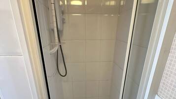 Ducha con mampara de vidrio, azulejos blancos, cabezal de ducha de mano y panel de control en la pared.