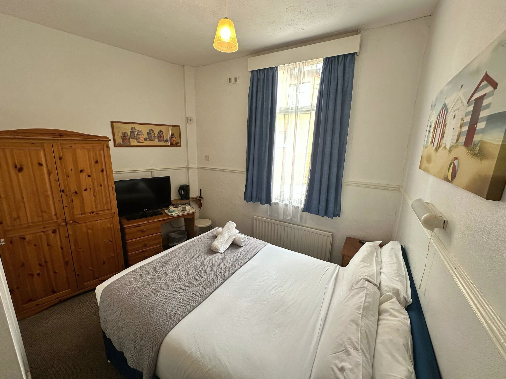 Une chambre  à coucher comprenant un lit, une armoire en bois, une télévision, un bureau avec un ordinateur et une fenêtre avec des rideaux.