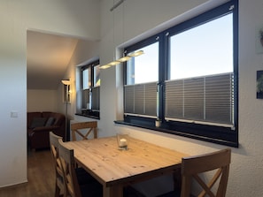 A dining area with a wooden table and chairs, a candle on the table, and a window with blinds. - Ferienwohnung in Burhave (Butjadingen)