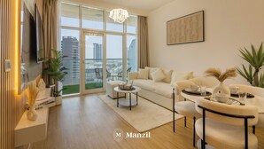 A modern living room with a sofa, coffee table, and dining area. A TV is mounted on the wall, and there is a large window with a city view. - Manzil -1BR w Pool nr Dubai Sports City  (Dubai)